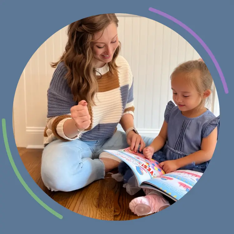 A smiling therapist teaching a young girl holding a book during an ABA therapy session.